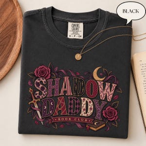 May include: A black short-sleeved t-shirt featuring a graphic print with the text SHADOW DADDY BOOK CLUB. The design includes a sword, roses, a crescent moon, and a book, with letters filled with various patterns in shades of purple and red.
