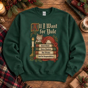 Może przedstawiać: Ciemnozielona bluza z okrągłym dekoltem z grafiką kota śpiącego na stosie książek. Tekst głosi All I Want for Yule, Good Books, Warm Blankets, Hot Drinks, And No Responsibilities. Przedstawiono również zapaloną świecę oraz gałązki ostrokrzewu.