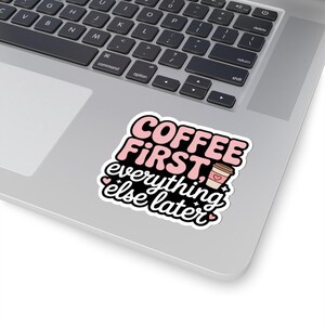 May include: A sticker with the text "COFFEE FIRST, everything else later" in pink and black. The sticker also features a coffee cup graphic with a heart. The sticker is on a laptop.