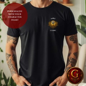 May include: A black short-sleeved t-shirt featuring a gold-colored emblem on the left chest. The emblem depicts a mace surrounded by ornate filigree, with the text SABLE CLERIC printed above and below the graphic in a white serif font.