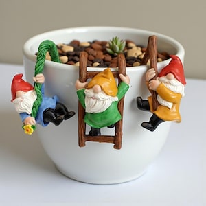 Könnte beinhalten: Ein weißer Keramik-Pflanztopf mit drei bunten Gartenzwergen. Ein Zwerg schwingt an einem grünen Seil, ein anderer klettert an einer Holzleiter und der dritte an einem Holzpfahl. Die Zwerge tragen rote, gelbe und orangefarbene Hüte und haben weiße Bärte.