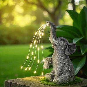 Pode incluir: Uma estátua de jardim de elefante cinzento com a tromba levantada, parecendo pulverizar um fluxo de luzes LED brancas quentes. O elefante está sentado numa base de pedra com musgo verde, contra um fundo verde desfocado.
