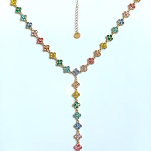 May include: A gold-tone Y-shaped necklace featuring a series of small, four-leaf clover-shaped charms. Each charm is encrusted with multi-coloured crystals in shades of pink, blue, green, yellow, and orange. The necklace includes a thin chain and a small circular tag.
