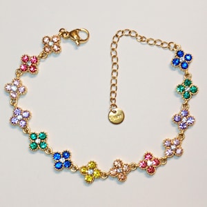 Könnte beinhalten: Ein goldfarbenes Kettenarmband mit mehreren kleeblattförmigen Anhängern, die jeweils mit kleinen, mehrfarbigen Kristallen in Rosa, Blau, Grün, Gelb und Violett besetzt sind. Es verfügt über einen Karabinerverschluss und ein kleines rundes Etikett.