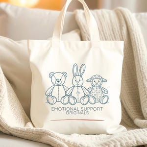 May include: Off-white tote bag with a line drawing of a teddy bear, a rabbit, and a lamb. The text "EMOTIONAL SUPPORT ORIGINALS" is printed below the illustrations. The bag has two handles.