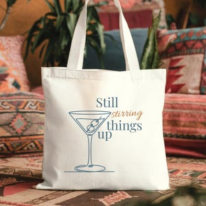 Könnte beinhalten: Eine weiße Canvas-Tragetasche mit dem Schriftzug "Still stirring things up" und einer Strichzeichnung eines Martiniglases. Das Martiniglas enthält Oliven. Die Tasche hat zwei weiße Griffe.