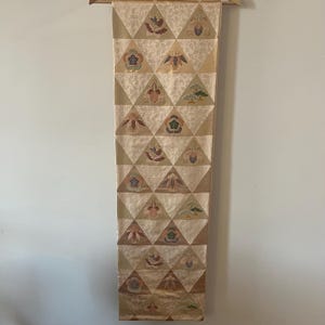 May include: A long, rectangular textile with a repeating pattern of embroidered triangles. The triangles are beige with floral and landscape motifs in various colors. The textile is hanging from a wooden rod.