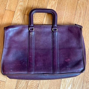 May include: A dark purple leather briefcase featuring a rectangular silhouette, a single top handle with a structured frame, and two vertical leather strips extending from the handle base to the bottom edge of the bag with a top zipper closure.
