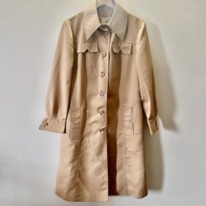 May include: A tan vintage trench coat with a large collar, button-up front, and pockets. The coat has long sleeves and a tailored fit, perfect for a retro look. The coat is made of a smooth, possibly satin-like material.