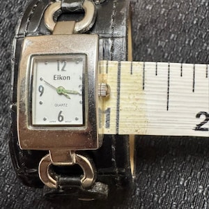 May include: A black leather cuff watch with a rectangular silver-toned face. The watch face displays the brand "Eikon", the time, and the word "Quartz". The watch band is approximately 2 inches wide.