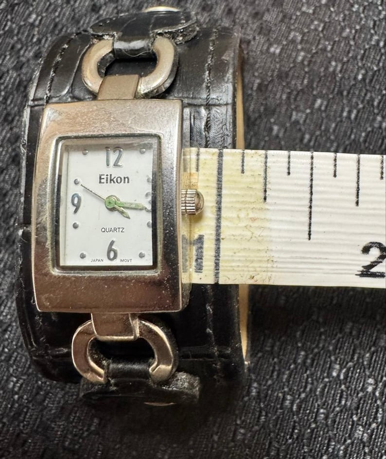 May include: A black leather cuff watch with a rectangular silver-toned face. The watch face displays the brand "Eikon", the time, and the word "Quartz". The watch band is approximately 2 inches wide.