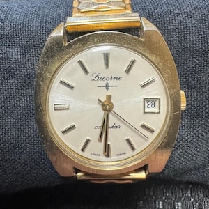 May include: Vintage Lucerne calendar watch with a gold-tone case and band. The watch has a white face with black markers, hands, and the date displayed at the 3 o'clock position. The watch face has the word "Lucerne" and "calendar" printed on it. The watch is marked "Swiss Made".