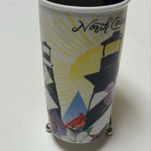 May include: White travel mug with a black lid, featuring a lighthouse design with black and white stripes, a yellow sun, and the text "North Carolina". Includes a red cardinal and floral accents.