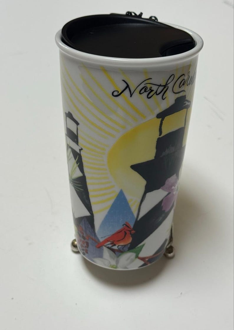 May include: White travel mug with a black lid, featuring a lighthouse design with black and white stripes, a yellow sun, and the text "North Carolina". Includes a red cardinal and floral accents.