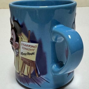 May include: A light blue ceramic mug featuring Mickey Mouse. The mug has a large handle and a design that includes Mickey Mouse and the text "Starring Tonight Mickey Mouse" on a small easel.