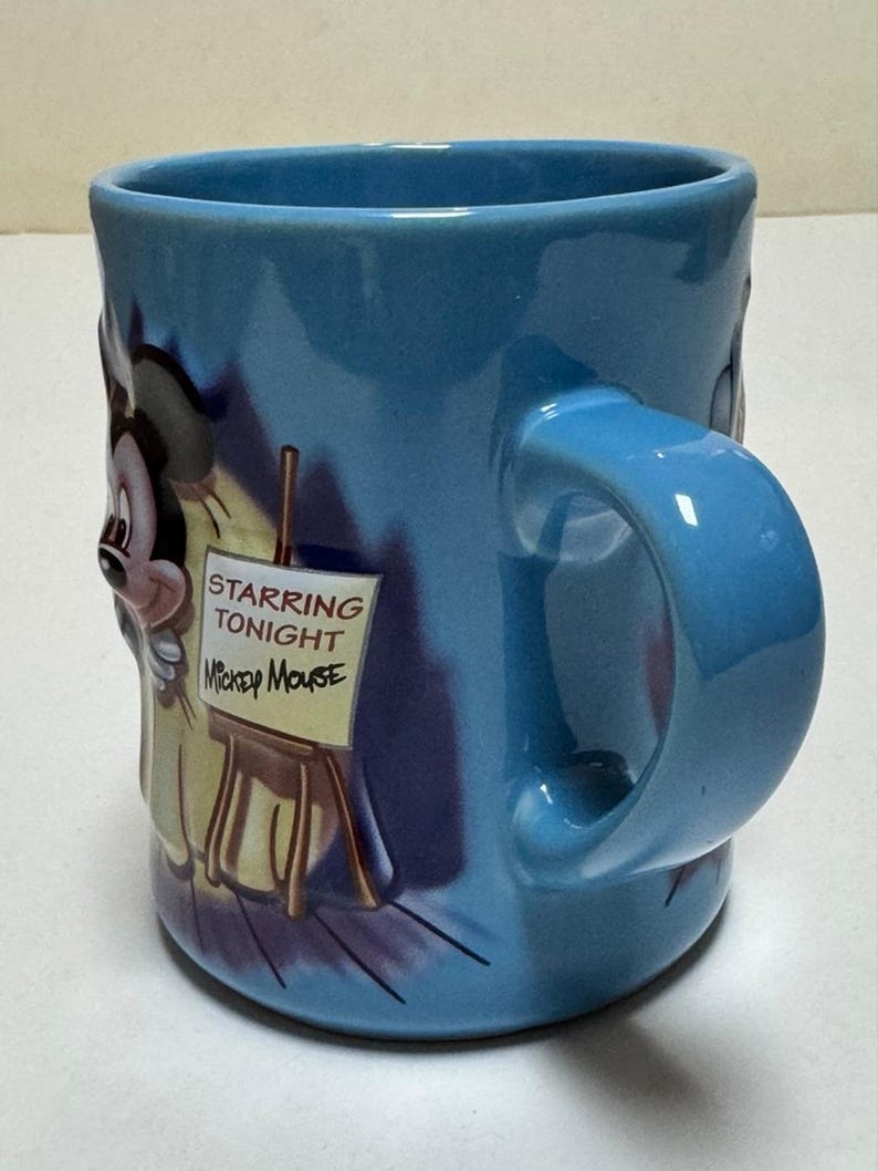 May include: A light blue ceramic mug featuring Mickey Mouse. The mug has a large handle and a design that includes Mickey Mouse and the text "Starring Tonight Mickey Mouse" on a small easel.