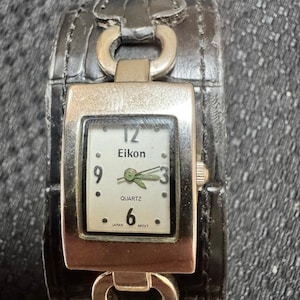 May include: Eikon quartz watch with a black leather cuff band. The watch face is square with a silver frame, white background, and black numerals. The watch band has silver metal accents.