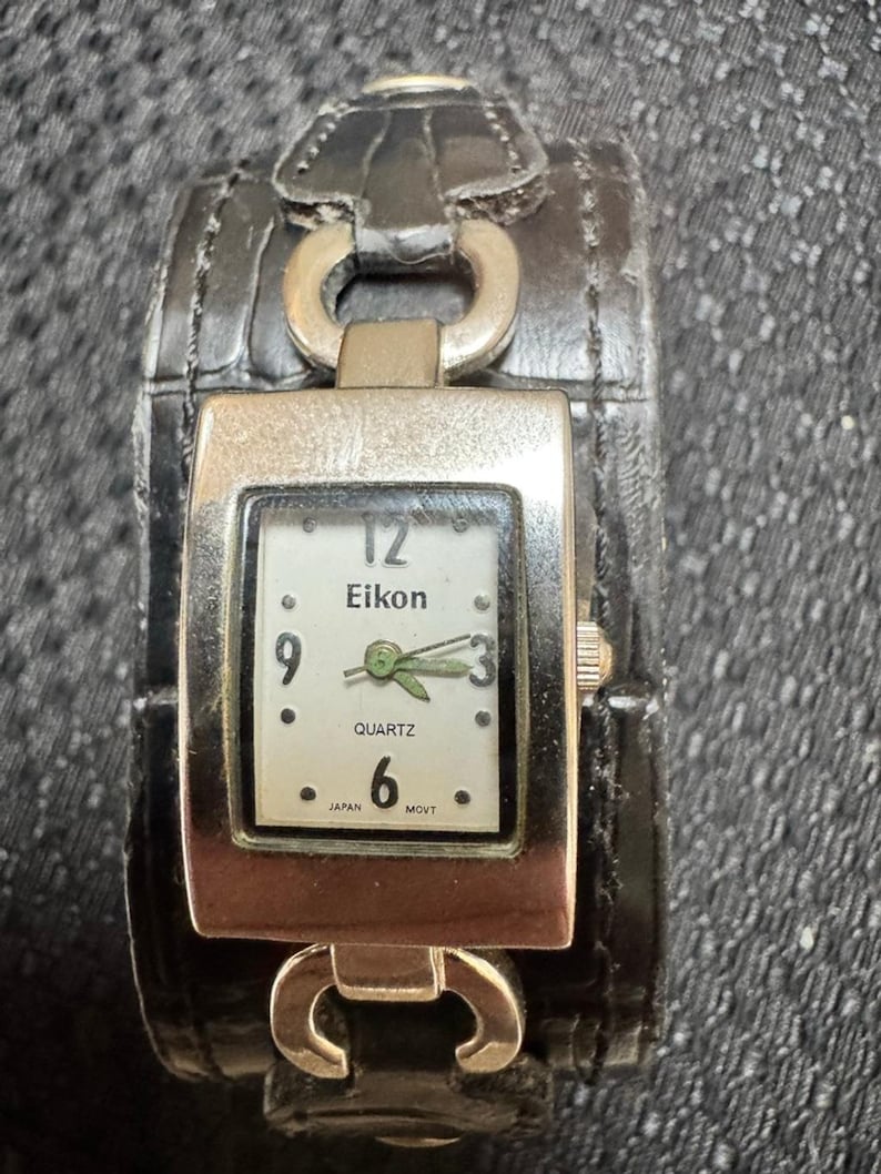 May include: Eikon quartz watch with a black leather cuff band. The watch face is square with a silver frame, white background, and black numerals. The watch band has silver metal accents.