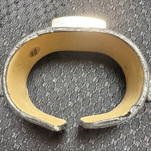 May include: A silver and black cuff bracelet with a rectangular silver accent. The bracelet has a textured, faux leather exterior and a light tan interior. The open design allows for easy wear.