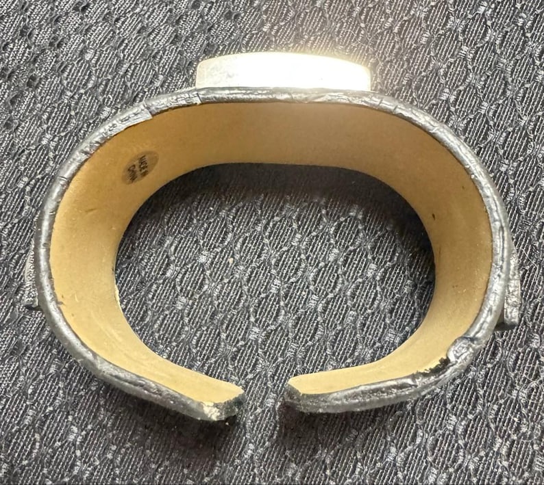 May include: A silver and black cuff bracelet with a rectangular silver accent. The bracelet has a textured, faux leather exterior and a light tan interior. The open design allows for easy wear.