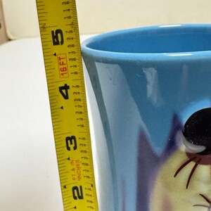 May include: A light blue ceramic mug with a cartoon character design. The mug is approximately 5 inches tall, as measured by a yellow tape measure. The mug has a glossy finish and a wide opening.
