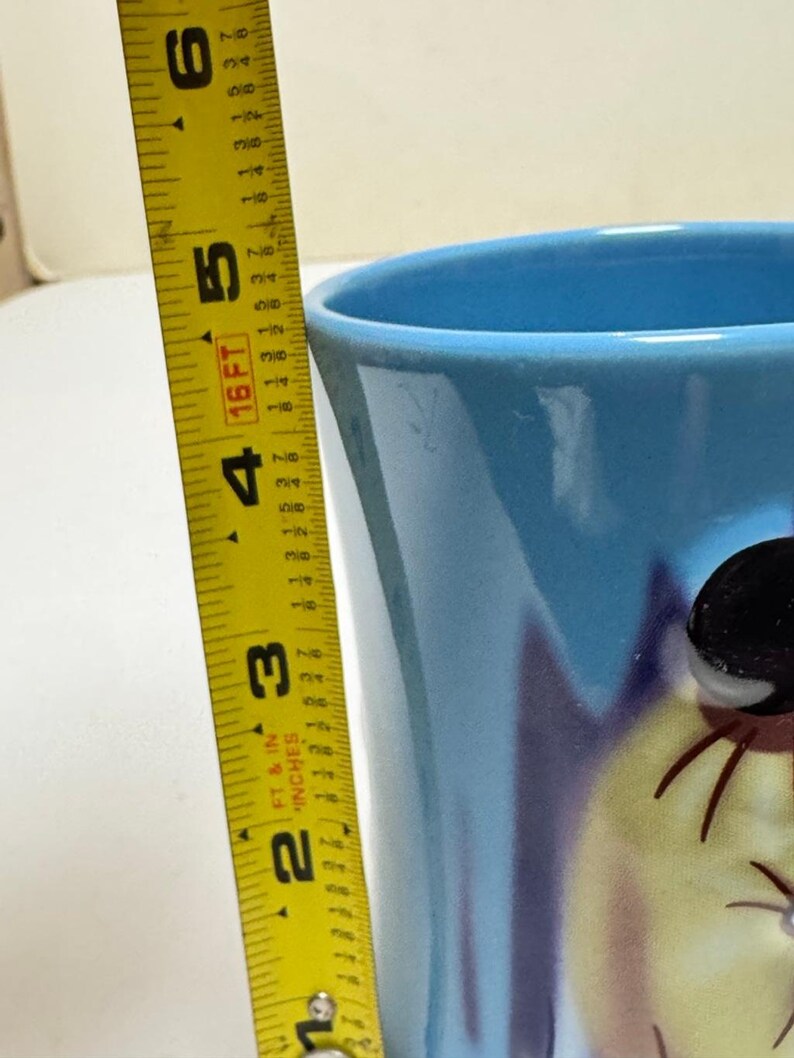 May include: A light blue ceramic mug with a cartoon character design. The mug is approximately 5 inches tall, as measured by a yellow tape measure. The mug has a glossy finish and a wide opening.