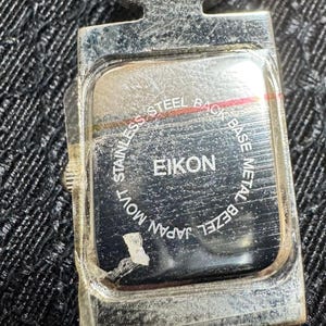 May include: Close-up of the back of a silver EIKON watch. The watch back is rectangular with rounded corners and features the text "STAINLESS STEEL BACK", "BASE METAL BEZEL", "JAPAN MOVT", and "EIKON".