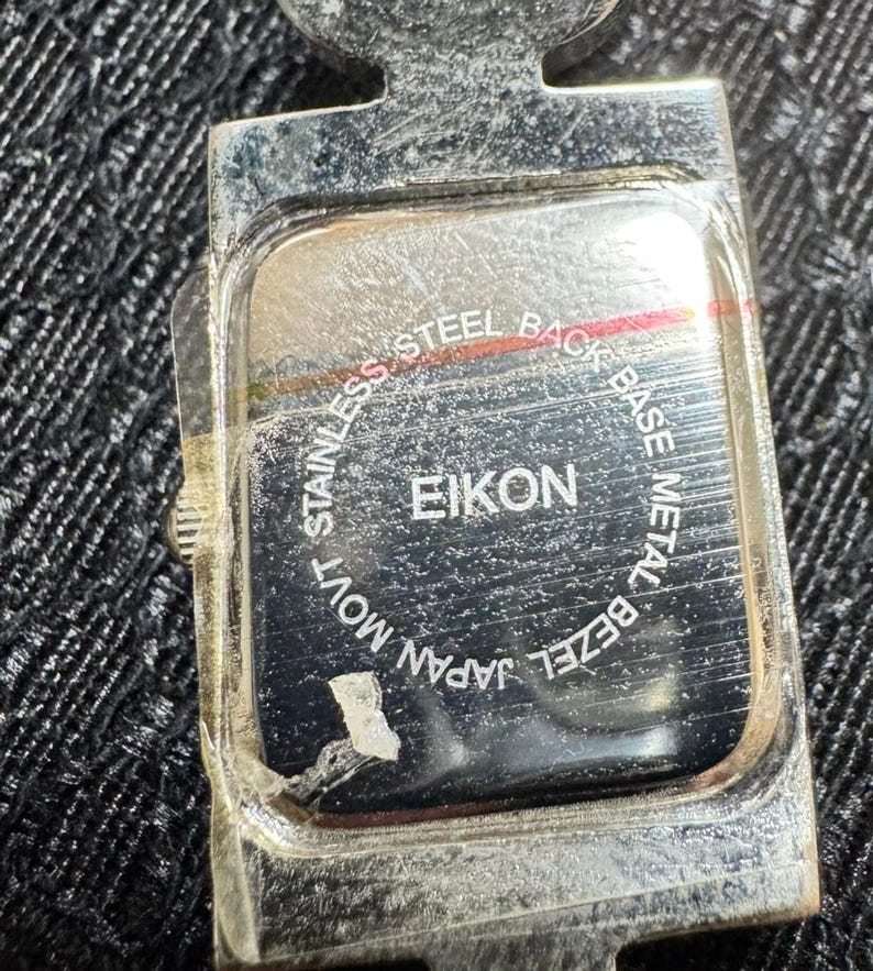 May include: Close-up of the back of a silver EIKON watch. The watch back is rectangular with rounded corners and features the text "STAINLESS STEEL BACK", "BASE METAL BEZEL", "JAPAN MOVT", and "EIKON".