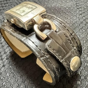 May include: A silver-toned watch with a rectangular face and a black leather cuff-style band. The watch face displays the time. The band features a buckle and a silver-toned button clasp. The watch is a stylish accessory.