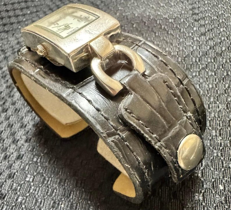 May include: A silver-toned watch with a rectangular face and a black leather cuff-style band. The watch face displays the time. The band features a buckle and a silver-toned button clasp. The watch is a stylish accessory.
