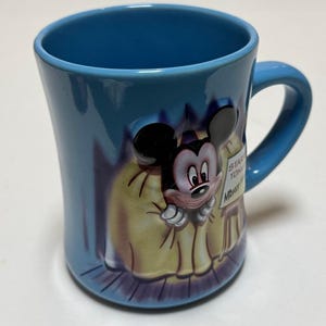 May include: A blue ceramic mug featuring a 3D relief of Mickey Mouse. The mug has a curved shape with a handle and a design of Mickey Mouse in a yellow coat. The mug has the words "STAR TON" and "Mickey" on a sign.
