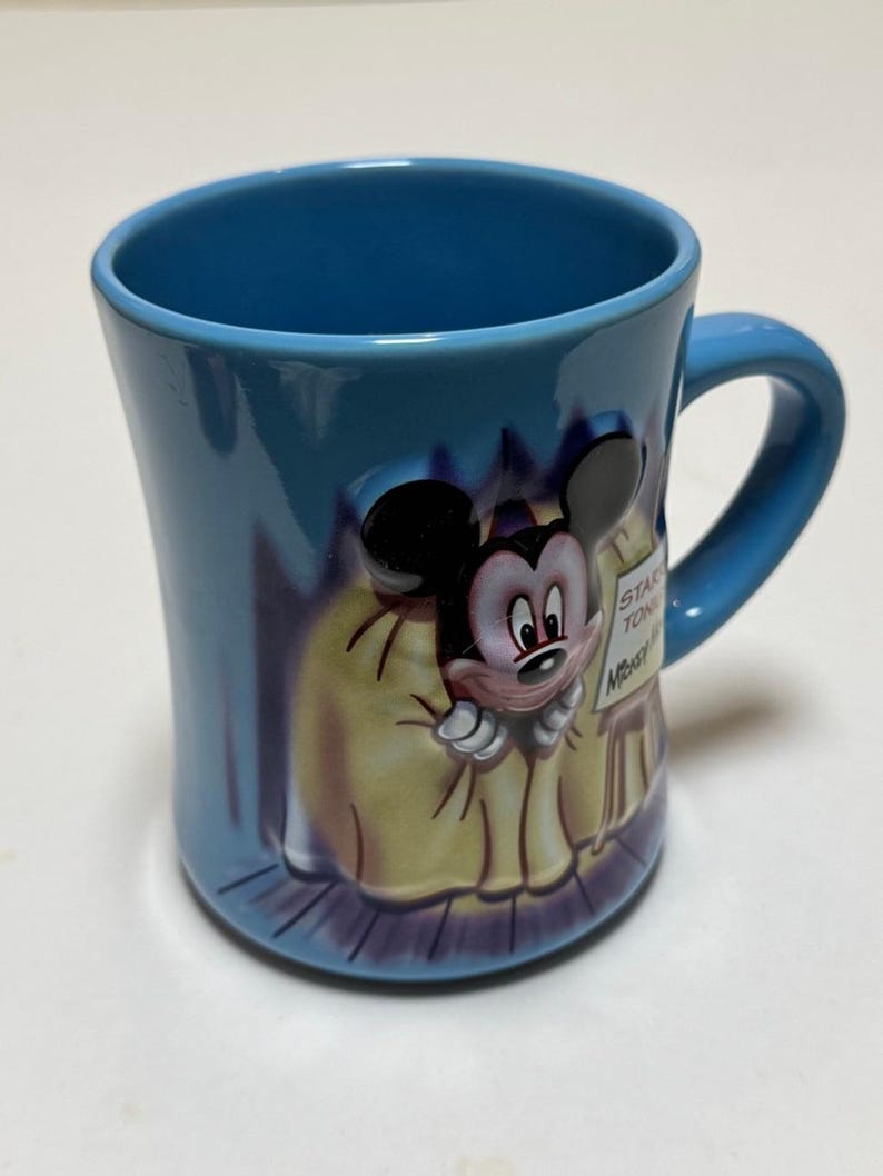 May include: A blue ceramic mug featuring a 3D relief of Mickey Mouse. The mug has a curved shape with a handle and a design of Mickey Mouse in a yellow coat. The mug has the words "STAR TON" and "Mickey" on a sign.