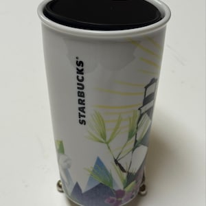 May include: White Starbucks travel mug with a black lid and a decorative design featuring a lighthouse, mountains, and floral elements. The mug has the Starbucks logo on the side.