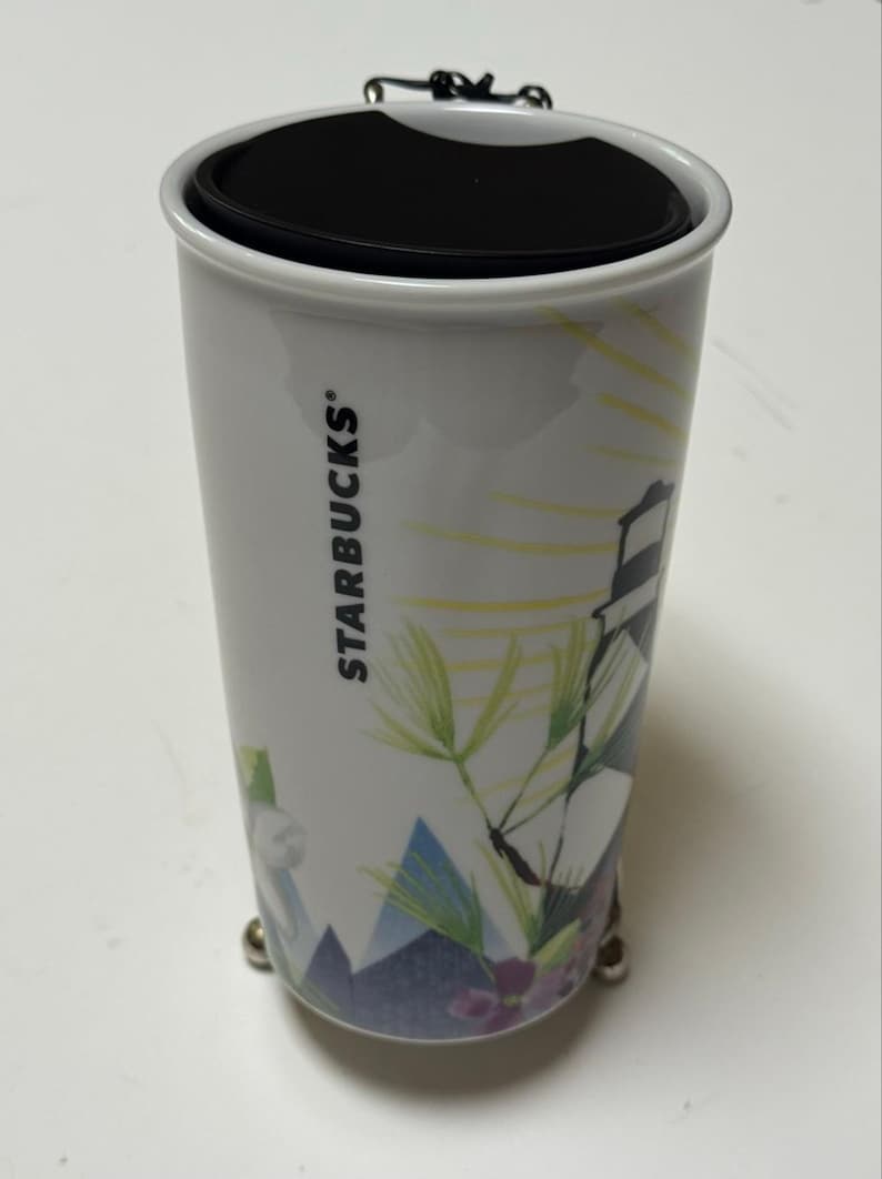 May include: White Starbucks travel mug with a black lid and a decorative design featuring a lighthouse, mountains, and floral elements. The mug has the Starbucks logo on the side.