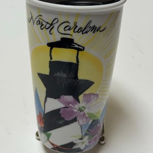 May include: White ceramic travel mug with a black lid, featuring a lighthouse design with black and white stripes, flowers, and the words "North Carolina". The mug has a yellow sunburst background.