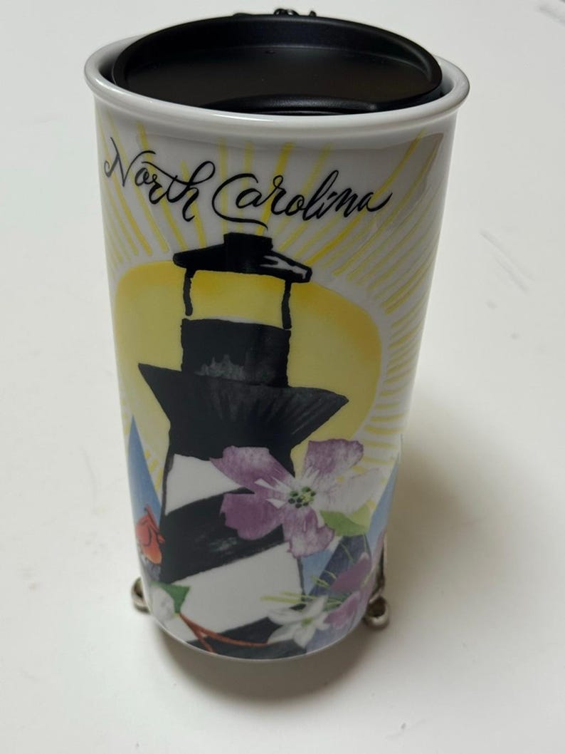 May include: White ceramic travel mug with a black lid, featuring a lighthouse design with black and white stripes, flowers, and the words "North Carolina". The mug has a yellow sunburst background.