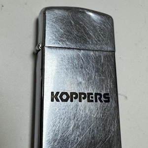 May include: A rectangular silver-toned metal lighter with a hinged lid and a brushed finish. The word KOPPERS is engraved in bold, dark capital letters on the front surface of the lighter body. The metal surface shows visible fine scratches.