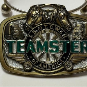 Puede incluir: Llavero vintage Teamster of America. El llavero presenta un emblema rectangular de color latón con las palabras "TEAMSTER OF AMERICA" en verde y blanco. El emblema tiene dos cabezas de caballo en la parte superior e imágenes de camiones a cada lado.
