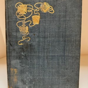 Könnte beinhalten: Ein dunkelgraues Hardcover-Buch mit einem goldfarbenen Prägemuster auf dem Vorderdeckel. Das Design zeigt ein seilartiges Muster, das ein Kreuz, ein Winkel- und Zirkelsymbol, einen Schild mit den Buchstaben K S T W S und einen Adler verbindet.