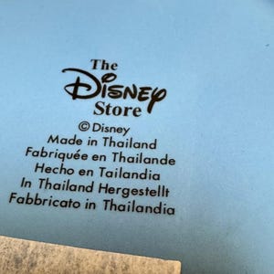 May include: A light blue ceramic plate with the text "The Disney Store" and "Made in Thailand" in multiple languages. The Disney logo is prominently displayed.
