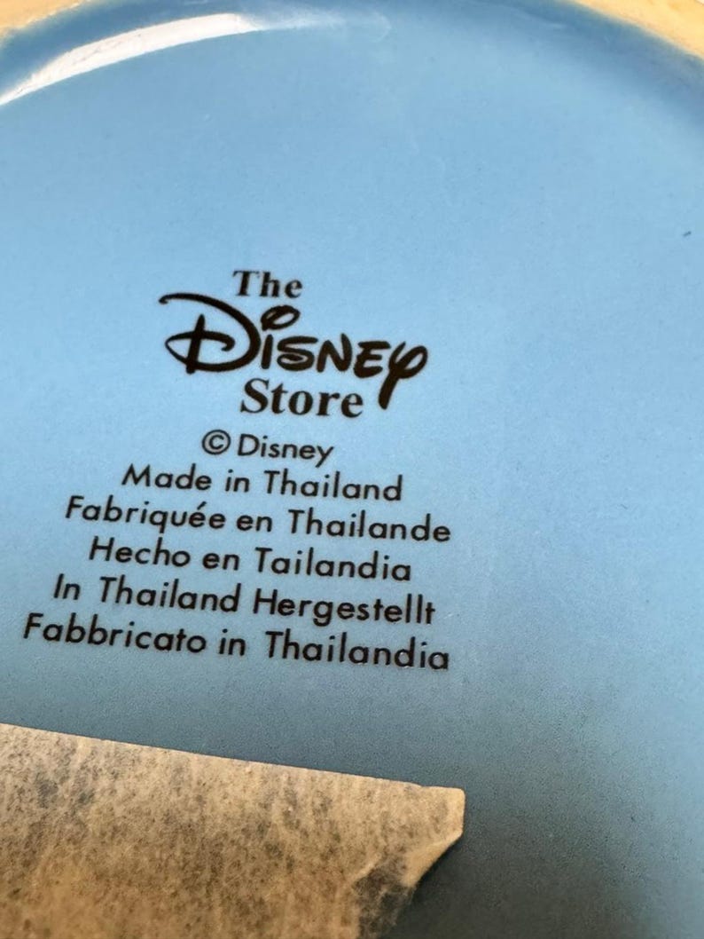 May include: A light blue ceramic plate with the text "The Disney Store" and "Made in Thailand" in multiple languages. The Disney logo is prominently displayed.