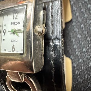 May include: A silver-toned Eikon Quartz watch with a rectangular face and a black leather band. The watch face is white with black numerals and green hands. The band is a wide cuff style.