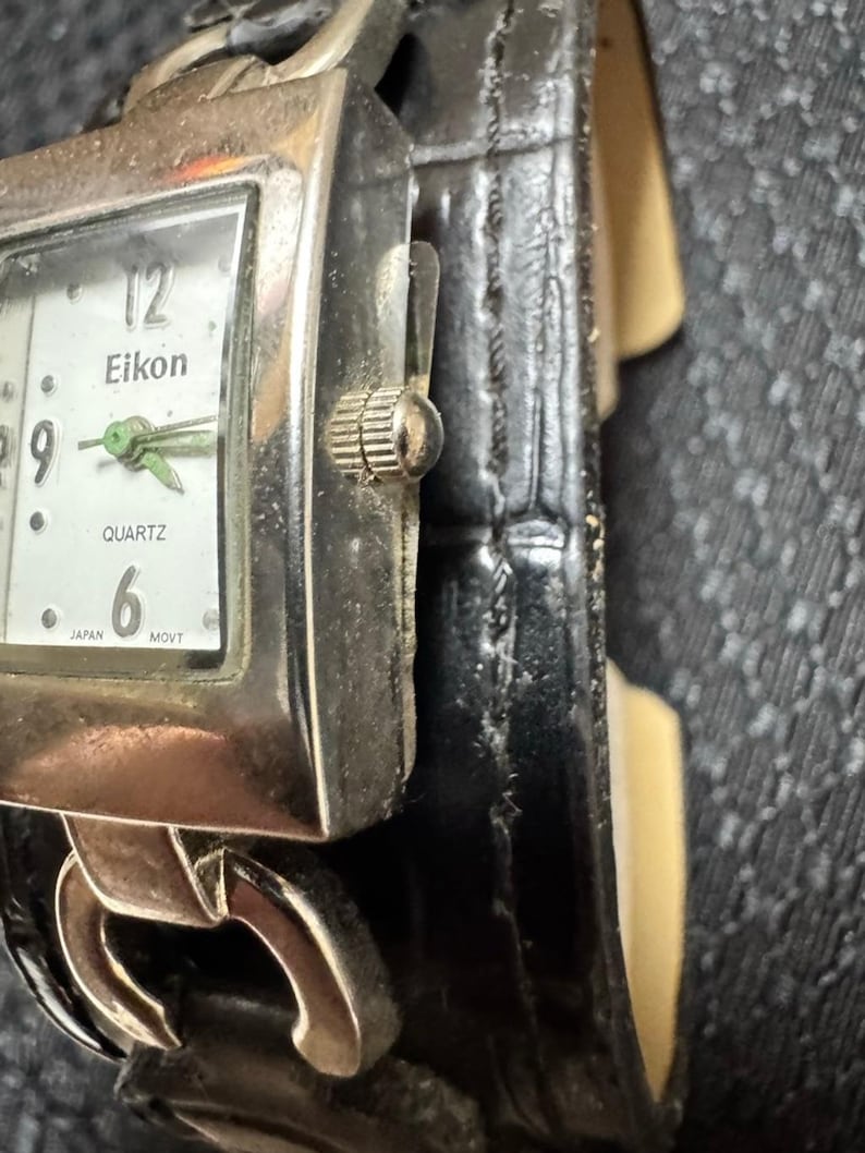 May include: A silver-toned Eikon Quartz watch with a rectangular face and a black leather band. The watch face is white with black numerals and green hands. The band is a wide cuff style.