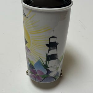 May include: White travel mug with a black lid and a silver handle. The mug features a colorful design of a lighthouse, sun, and flowers. The mug is cylindrical and has a black lid.