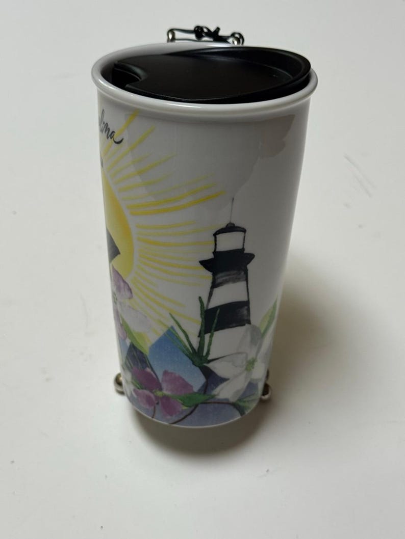 May include: White travel mug with a black lid and a silver handle. The mug features a colorful design of a lighthouse, sun, and flowers. The mug is cylindrical and has a black lid.