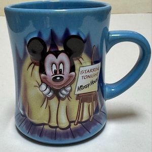 May include: A blue ceramic mug featuring Mickey Mouse. The mug has a curved handle and a design of Mickey Mouse on a stage with the text "Starring Tonight Mickey Mouse" on a small easel. The mug is a light blue color.