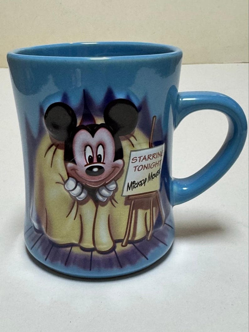 May include: A blue ceramic mug featuring Mickey Mouse. The mug has a curved handle and a design of Mickey Mouse on a stage with the text "Starring Tonight Mickey Mouse" on a small easel. The mug is a light blue color.