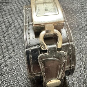 May include: A silver Eikon watch with a rectangular face and a black leather band. The watch face displays the brand name and the time. The band is wide and has a textured pattern.