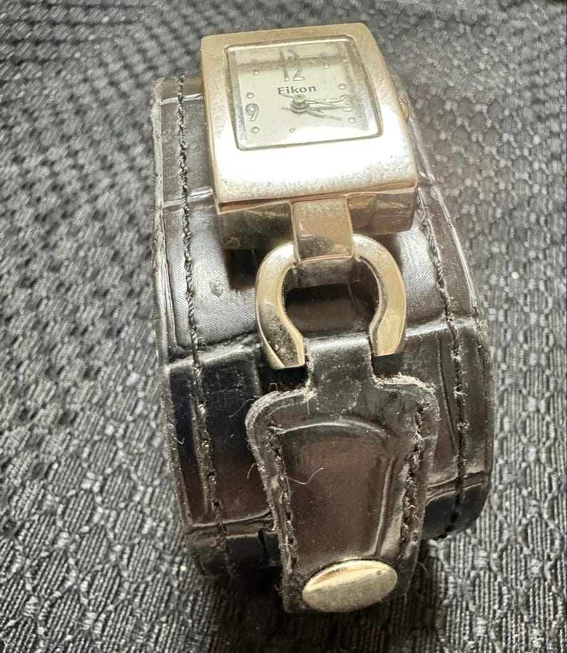 May include: A silver Eikon watch with a rectangular face and a black leather band. The watch face displays the brand name and the time. The band is wide and has a textured pattern.