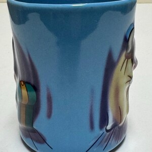 May include: A blue ceramic mug with a cartoon-style design. The mug features a light blue exterior with a darker blue and purple design on the sides. The design includes a cartoon face and a small, colorful object.